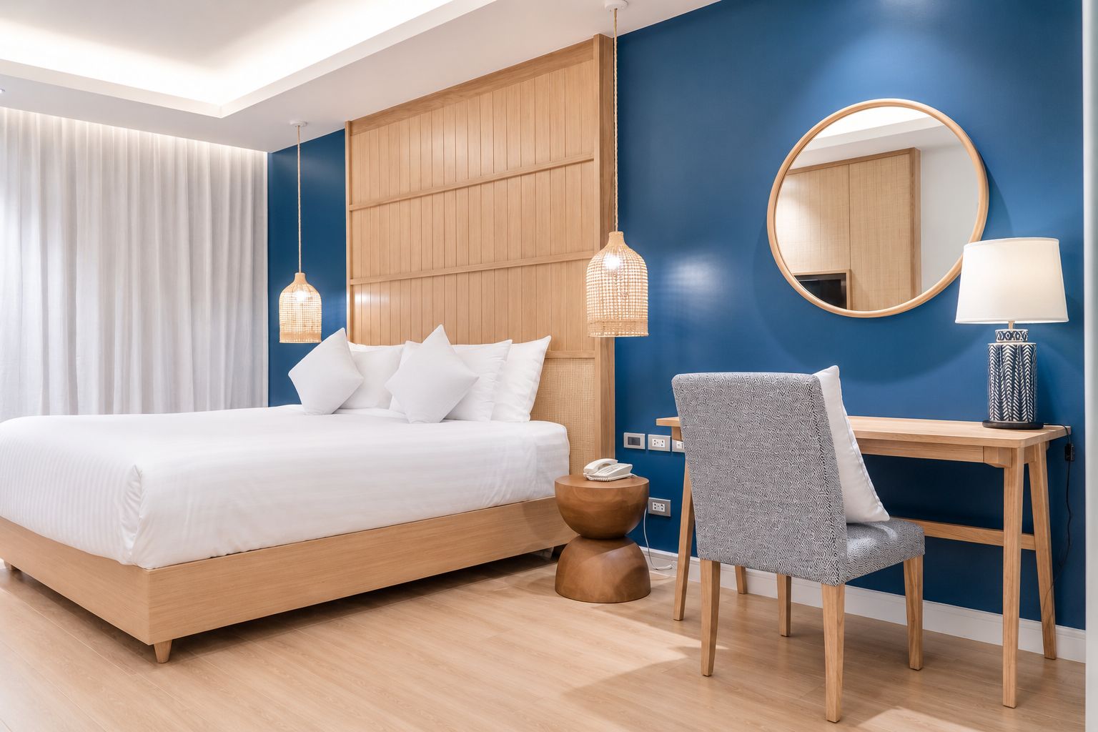 A hotel room with a bed, a desk, a chair, a round mirror, and a hanging light fixture.