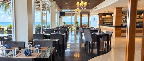 Un restaurant modern amb vista a l'oceà, que destaca per una àrea de menjador ampla amb taules i cadires, una zona de bar i un sostre de fusta.