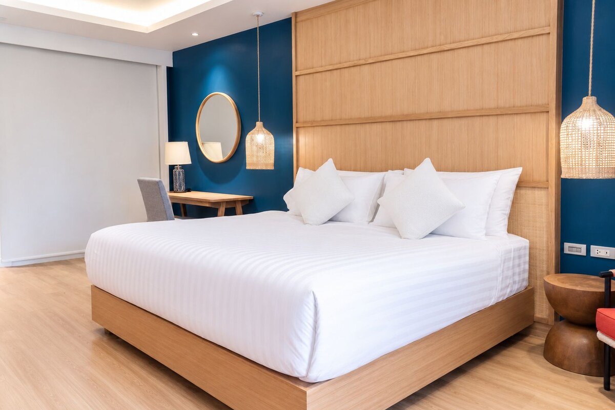 Una habitación de hotel moderna con una cama grande, un escritorio y una silla. Hay un espejo en la pared y una lámpara colgante.