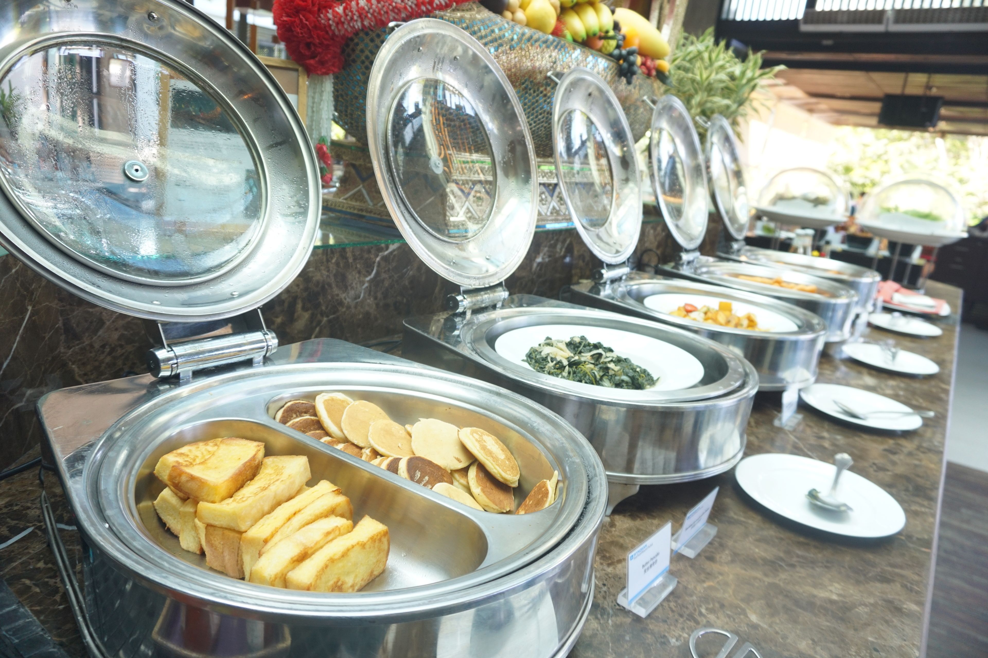 Buffet com diversos alimentos em recipientes metálicos, incluindo panquecas e um prato com vegetais.