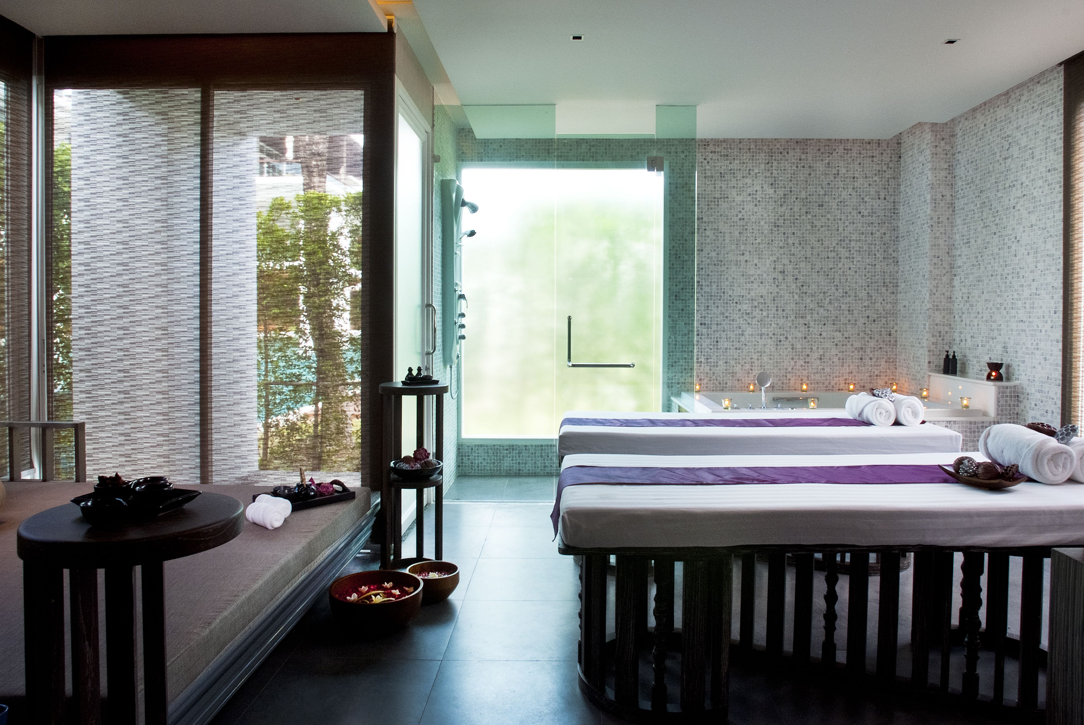 A modern hotel room with a large window, two beds, a small table, and a bathroom visible through the glass door.