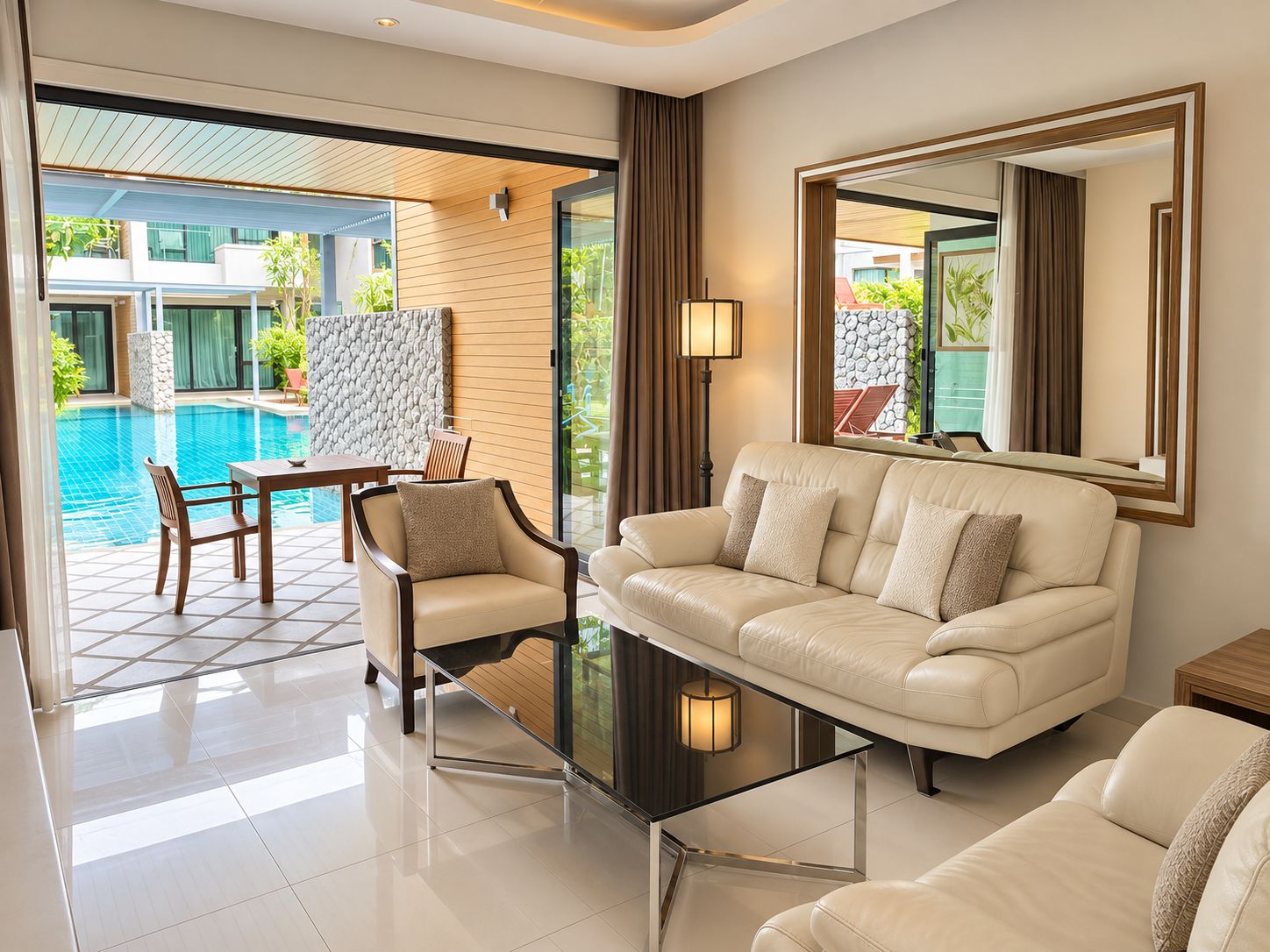 A modern living room with a sofa, armchairs, and a coffee table. There is a view of a swimming pool and outdoor furniture.