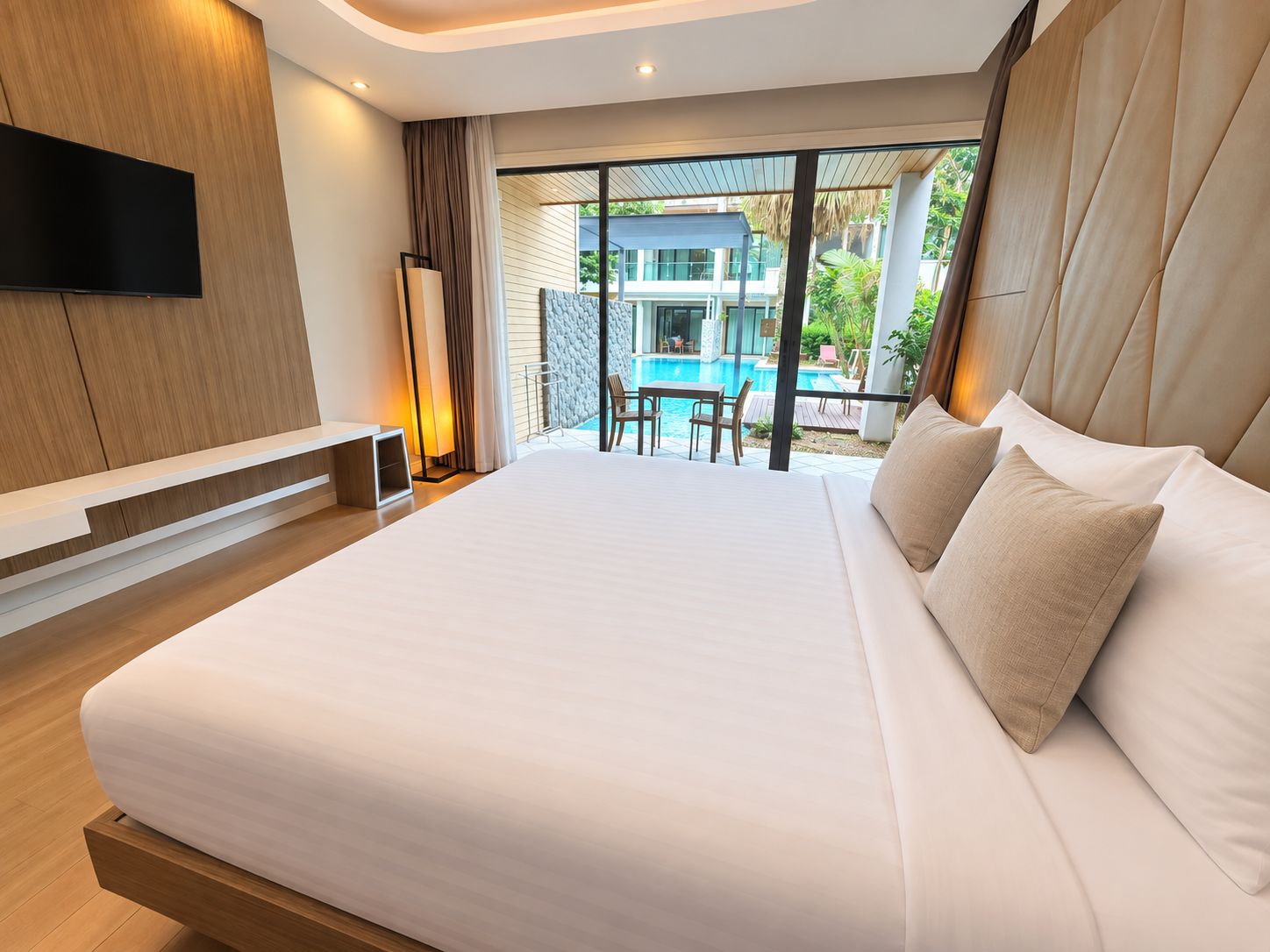 A modern hotel room with a large bed, a flat-screen TV, and a view of a swimming pool and outdoor seating.
