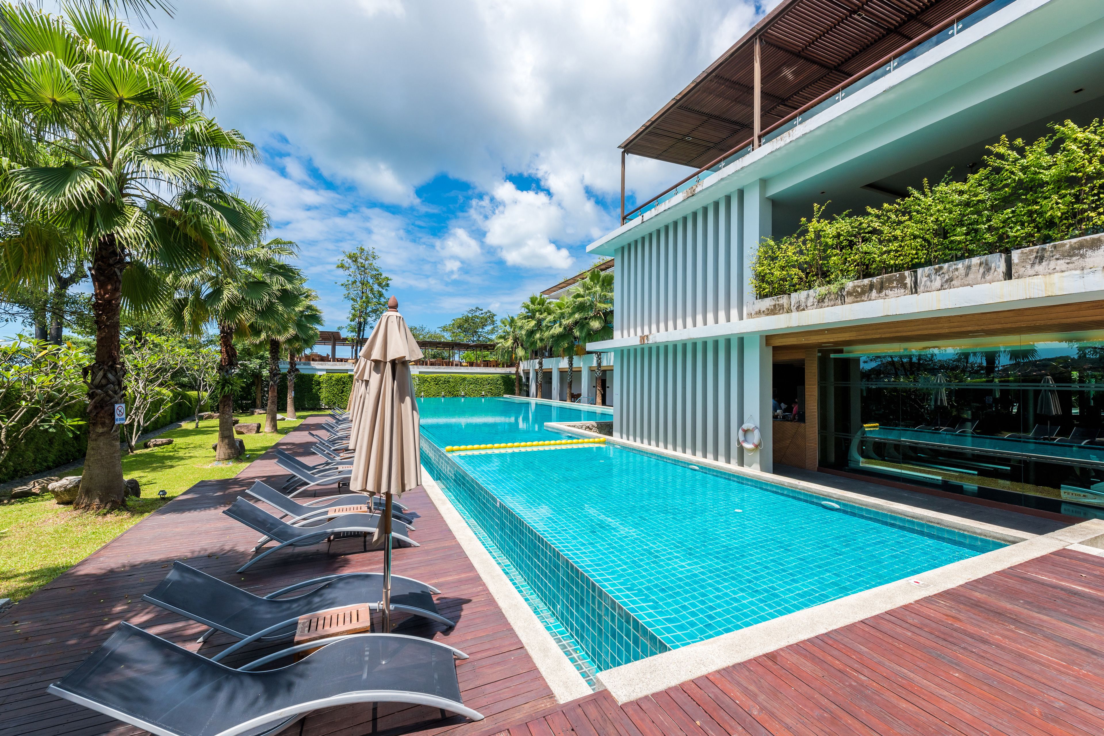 A swimming pool with lounge chairs and umbrellas, surrounded by greenery and a building with a balcony.
