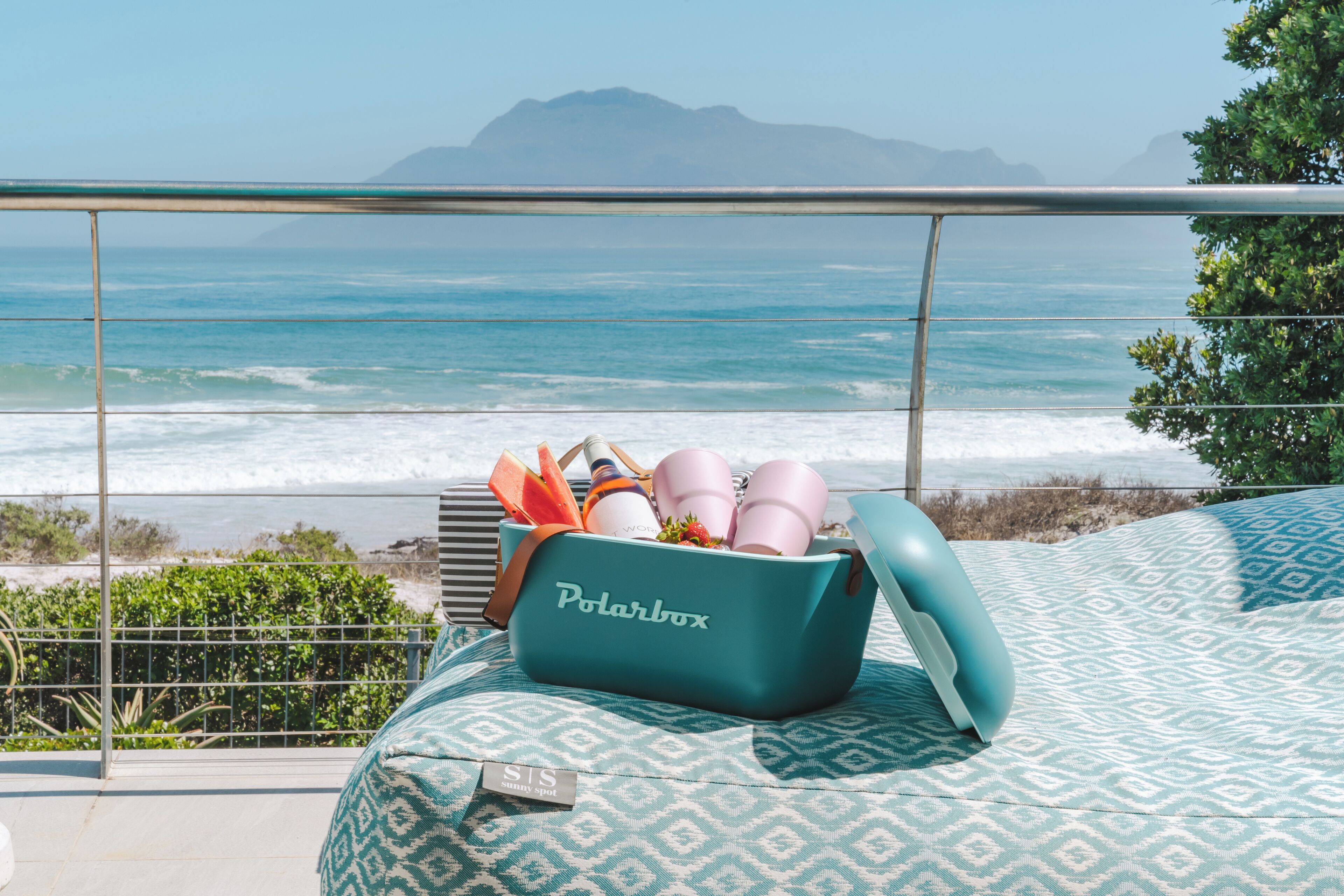 Una bolsa color turquesa con la etiqueta "Polarbox" sobre una cama estampada, con vista al océano y montañas al fondo.