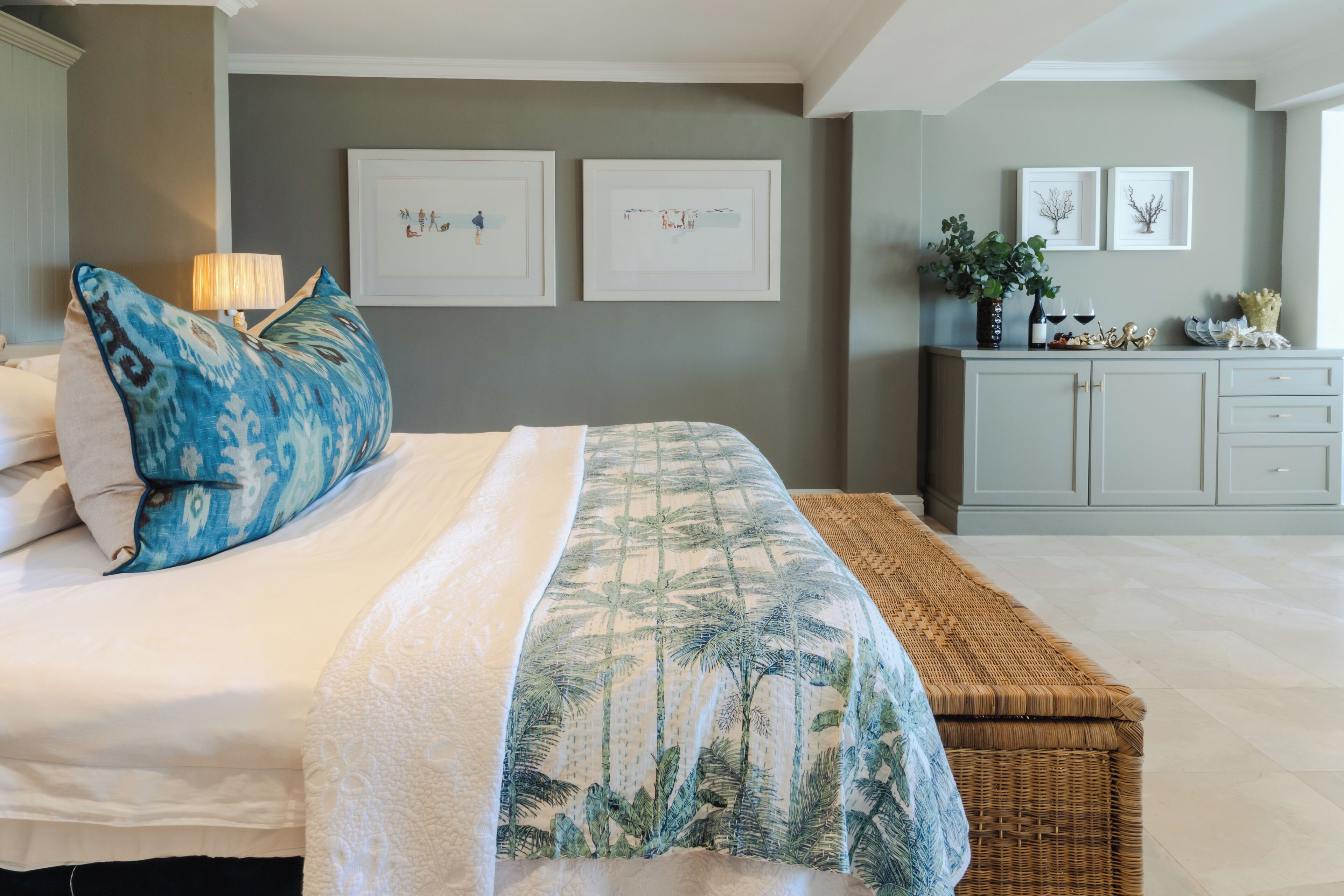 A bedroom with a bed, a bedside table with a lamp, a wicker bench, and a wall with framed pictures.
