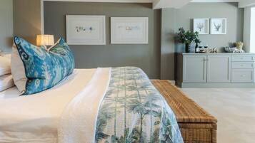 A bedroom with a bed, a bedside table with a lamp, a wicker bench, and a wall with framed pictures.