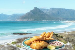 A plate of fish and chips with a side of tartar sauce and lemon wedges, set against a coastal backdrop with mountains. - Last Word Long Beach (Cape Town)