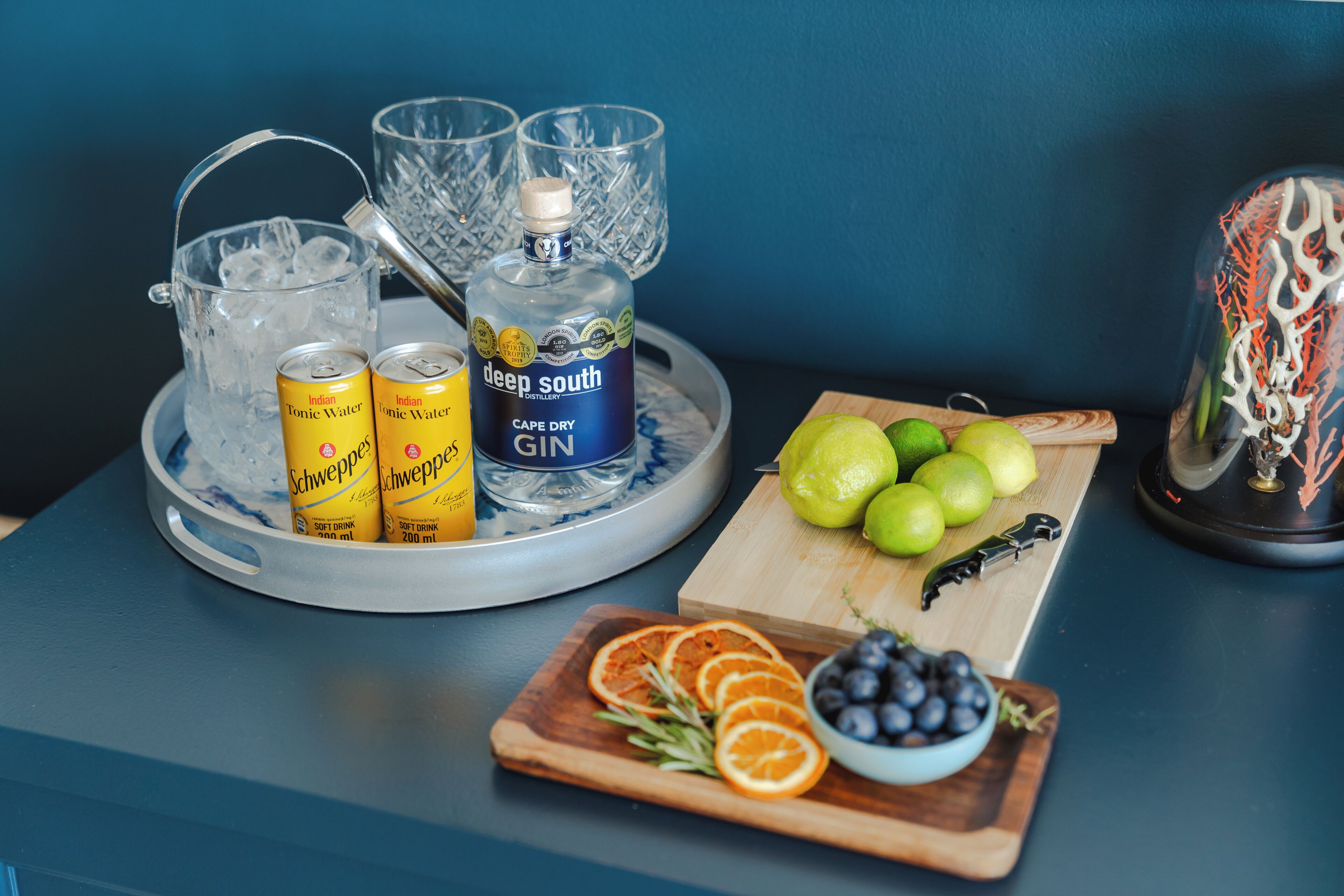 En bakke med tonicvand, gin og glas, et skærebræt med limefrugter og en kniv, samt en skål med blåbær og appelsiner.