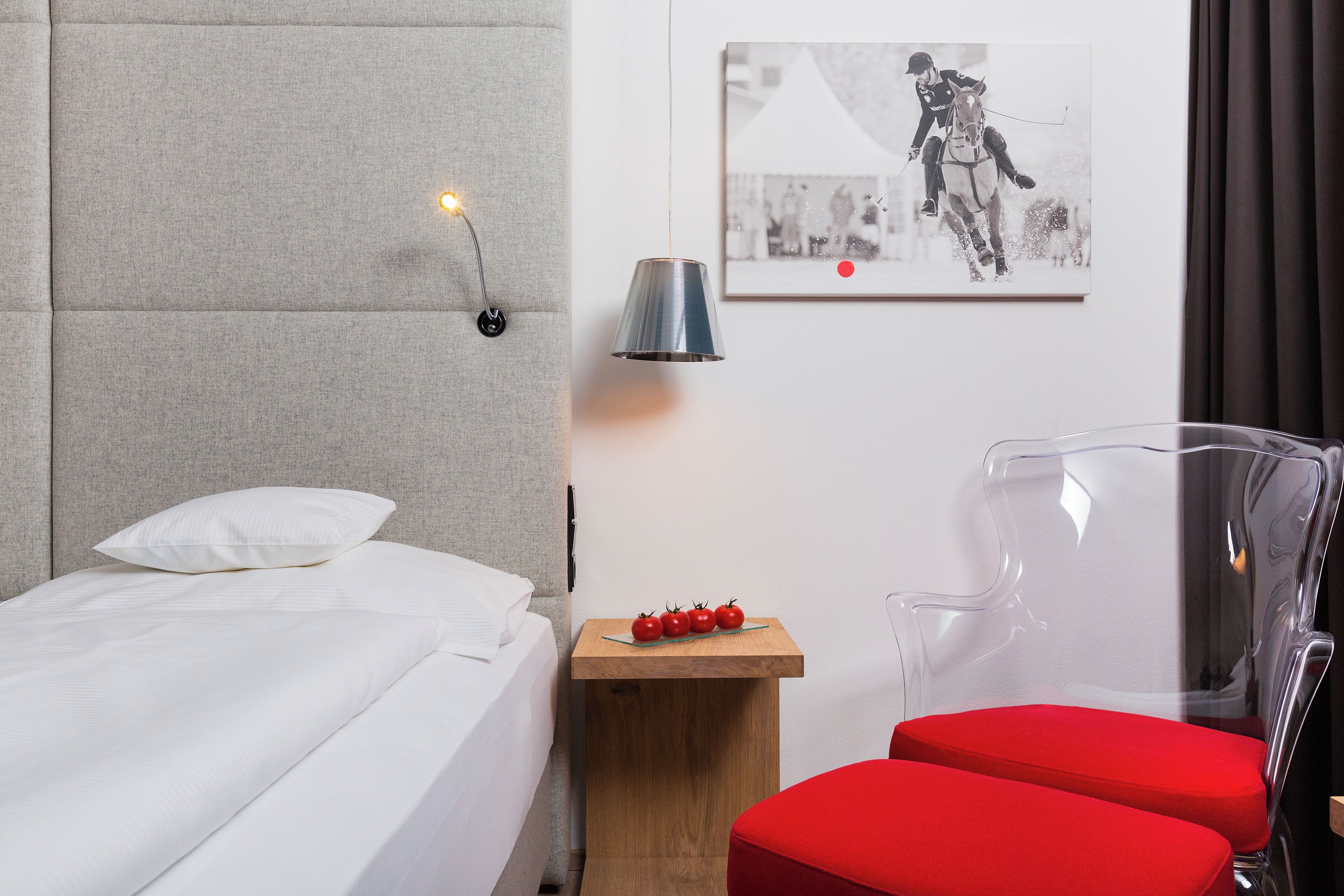 A hotel room with a bed, a side table with a lamp, a small wooden table with tomatoes, and a red chair.