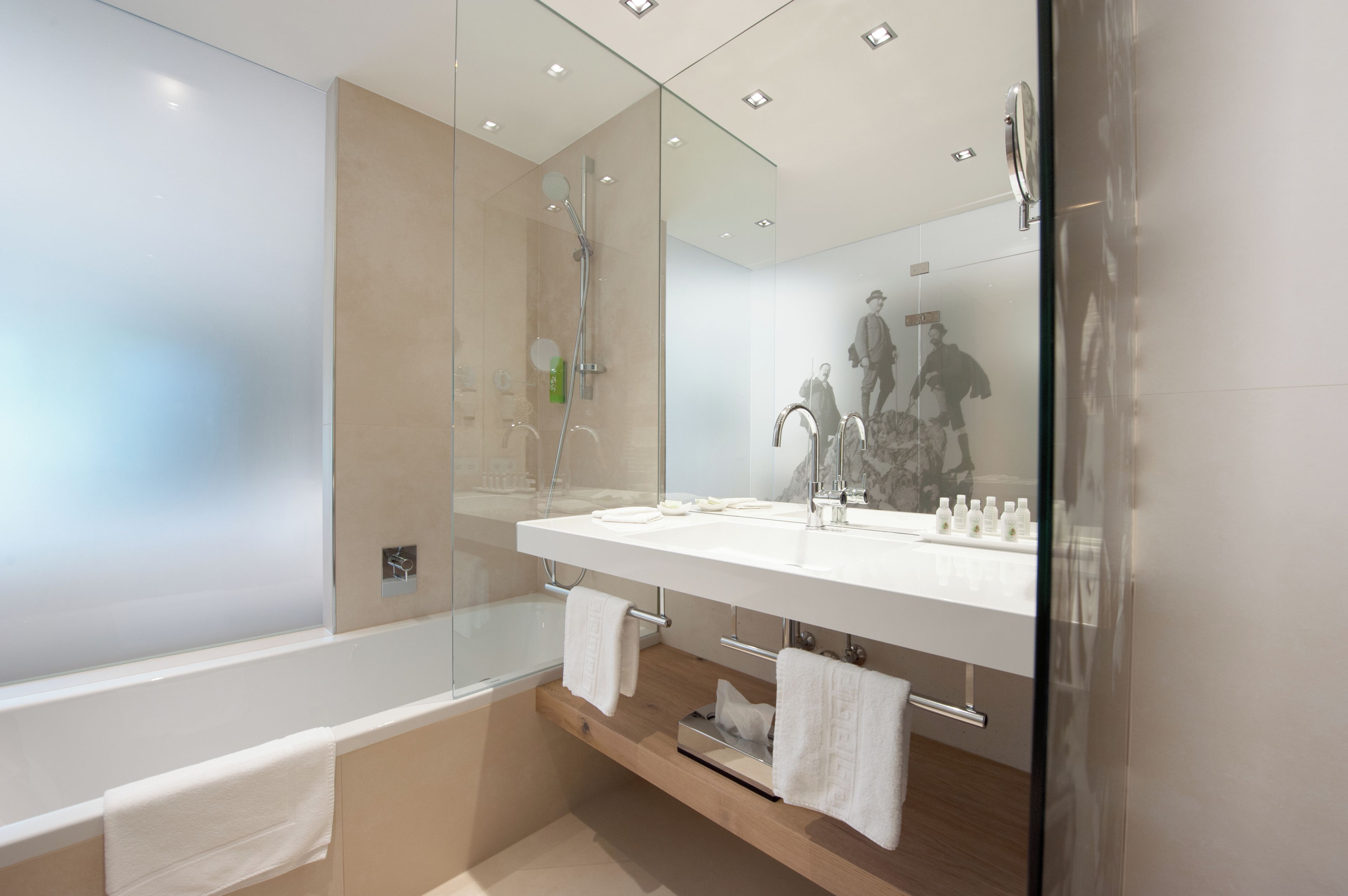 A modern bathroom with a glass-enclosed shower, a white bathtub, and a large mirror reflecting a person.