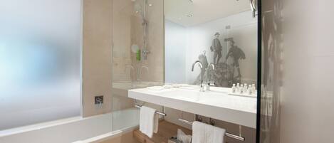A modern bathroom with a glass-enclosed shower, a white bathtub, and a large mirror reflecting a person.