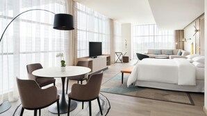 Een moderne hotelkamer met een groot bed, een televisie, een zithoek met een tafel en stoelen, en grote ramen.