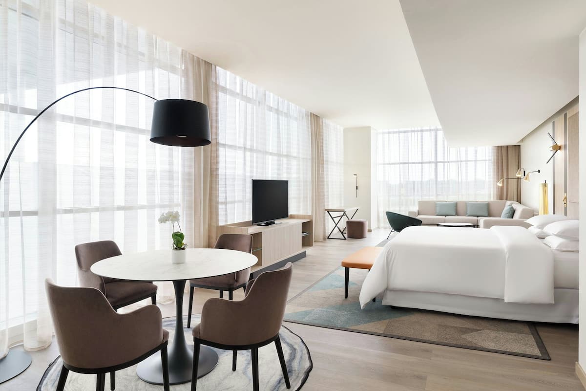 Ein modernes Hotelzimmer mit einem großen Bett, einem Fernseher, einem Essbereich mit Tisch und Stühlen sowie großen Fenstern.