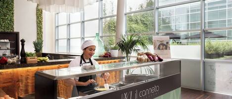 A modern restaurant interior with a chef preparing food at a counter, large windows, and a green wall.