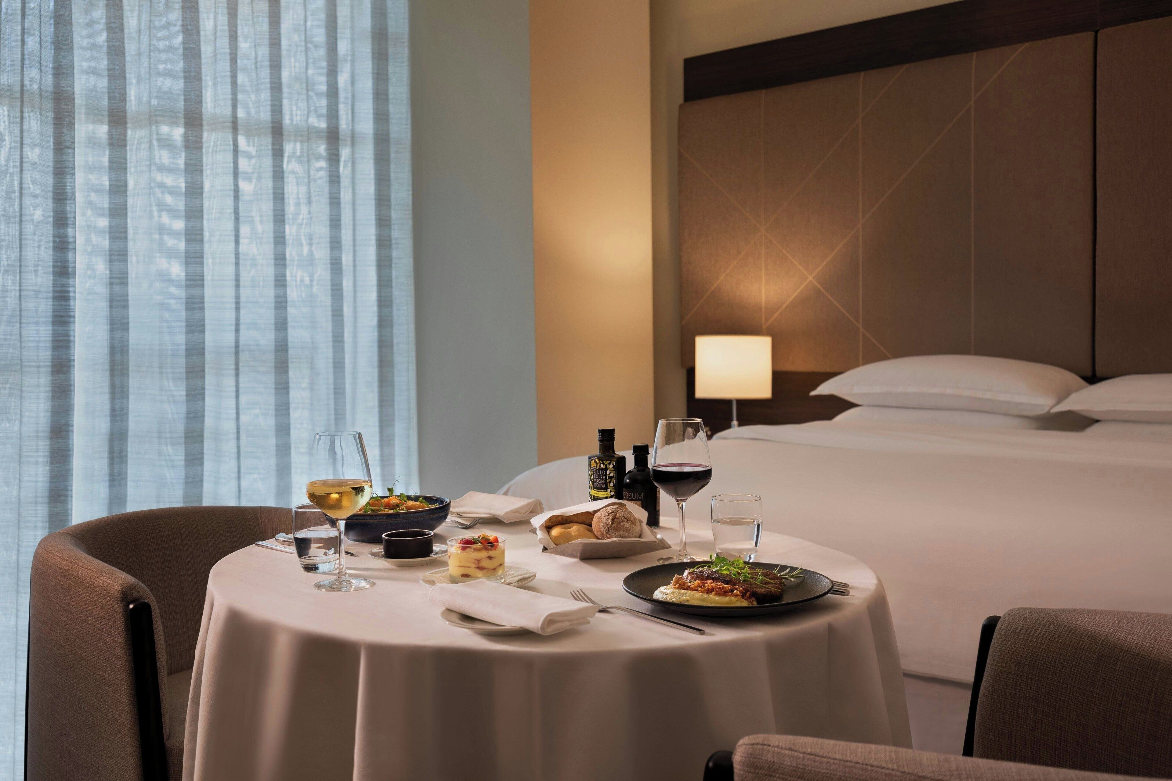 A hotel room with a dining table set for a meal, a bed with pillows, and a nightstand with a lamp.