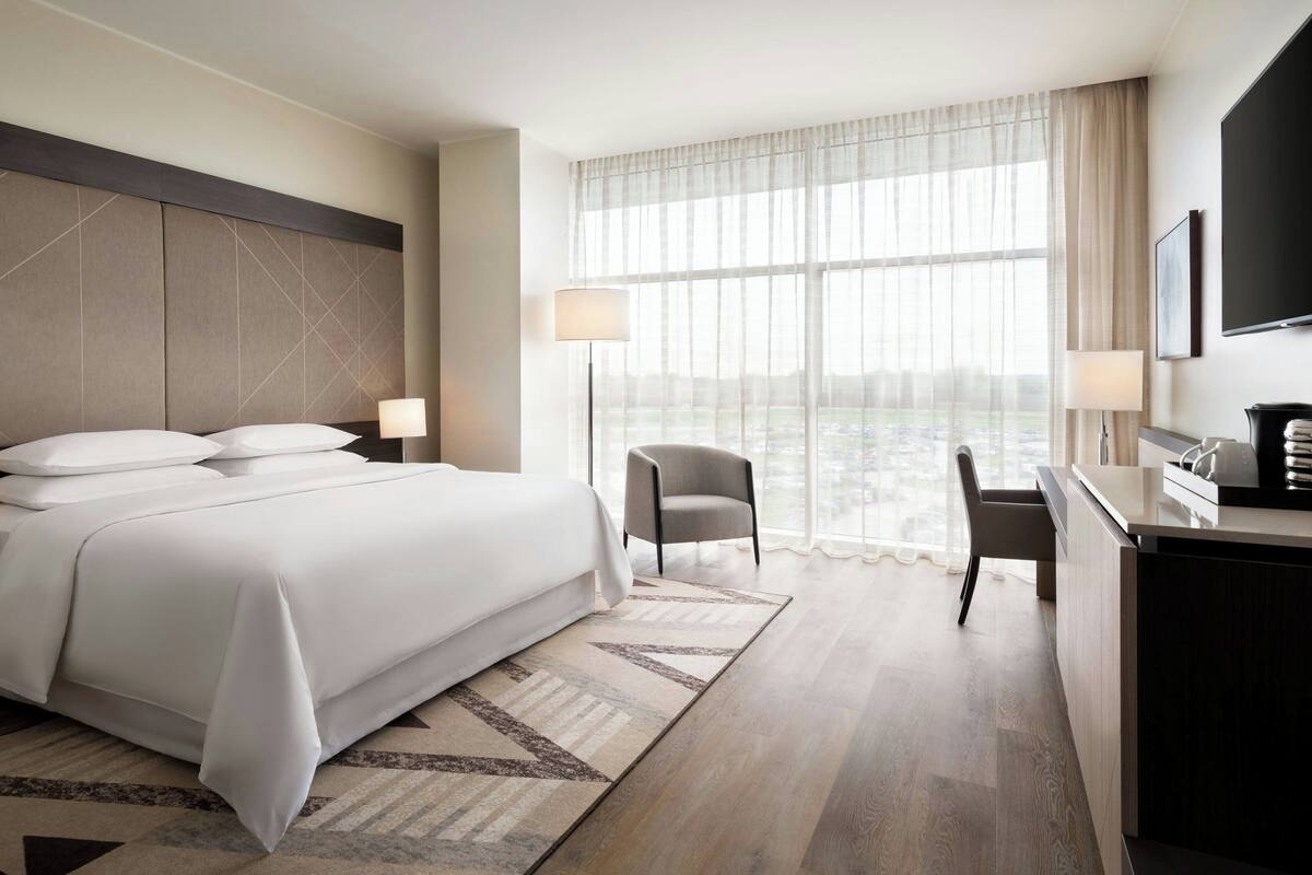 Ein Hotelzimmer mit einem großen Bett, einem Schreibtisch, einem Sessel, einem Fernseher und Blick auf die Stadt durch große Fenster.