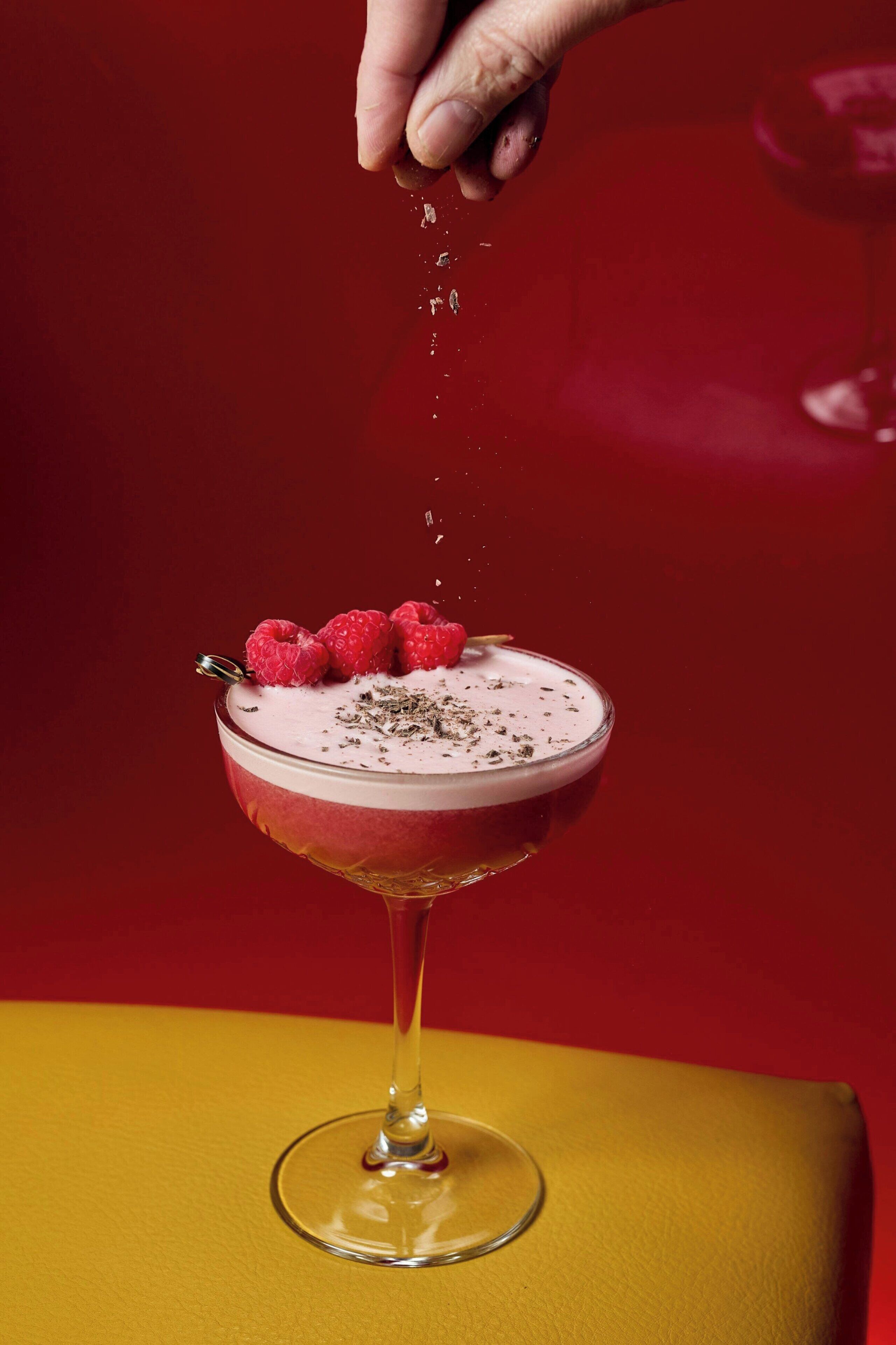 A cocktail glass with a layered drink, garnished with raspberries and a sprinkle of herbs, on a yellow surface.
