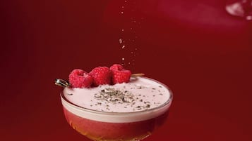 Un vaso de cóctel con una bebida en capas, adornado con frambuesas y una pizca de hierbas, sobre una superficie amarilla.