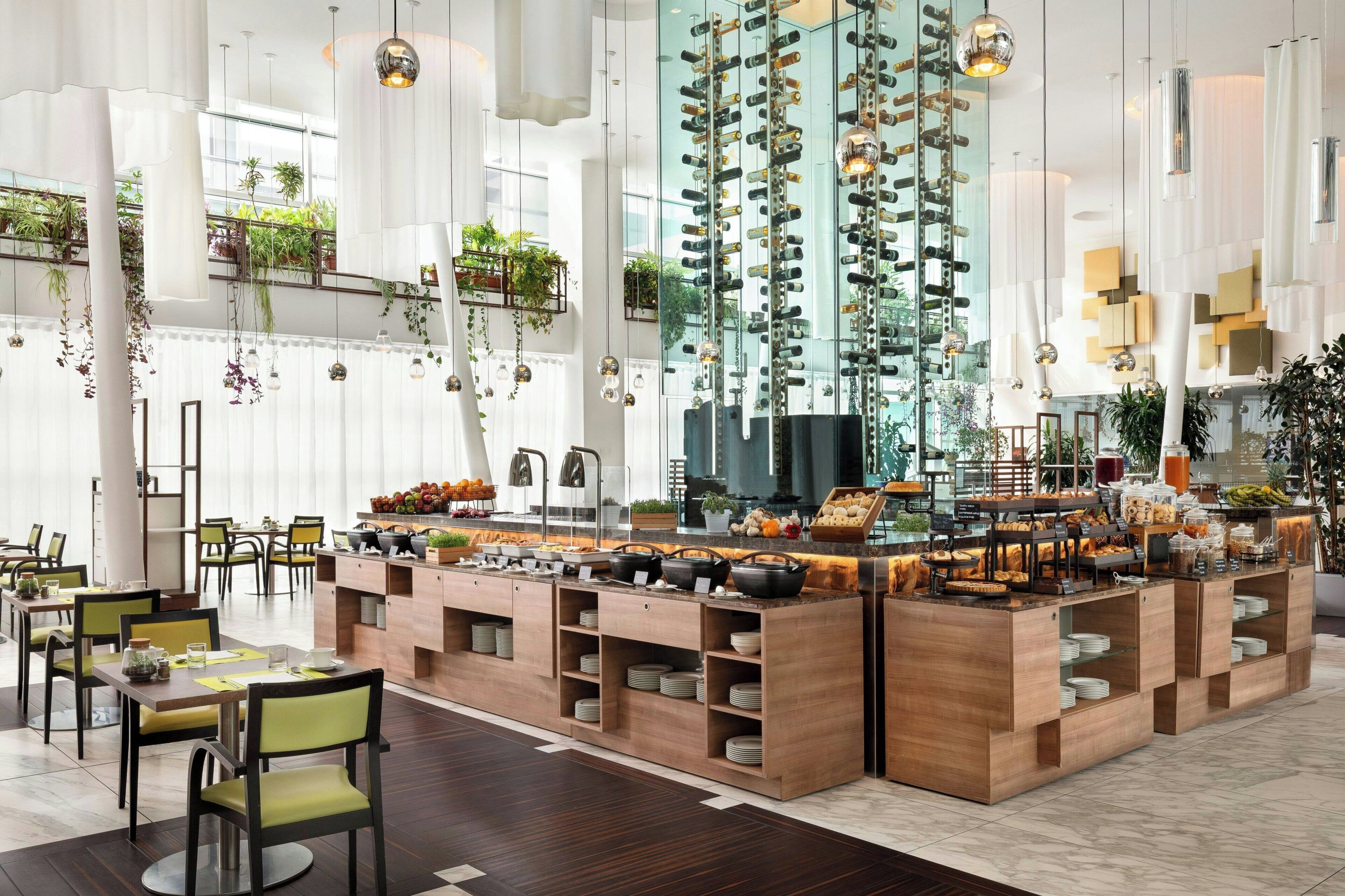 Un comedor moderno con mobiliario de madera, un bufé central con diversas opciones de comida y plantas colgantes.