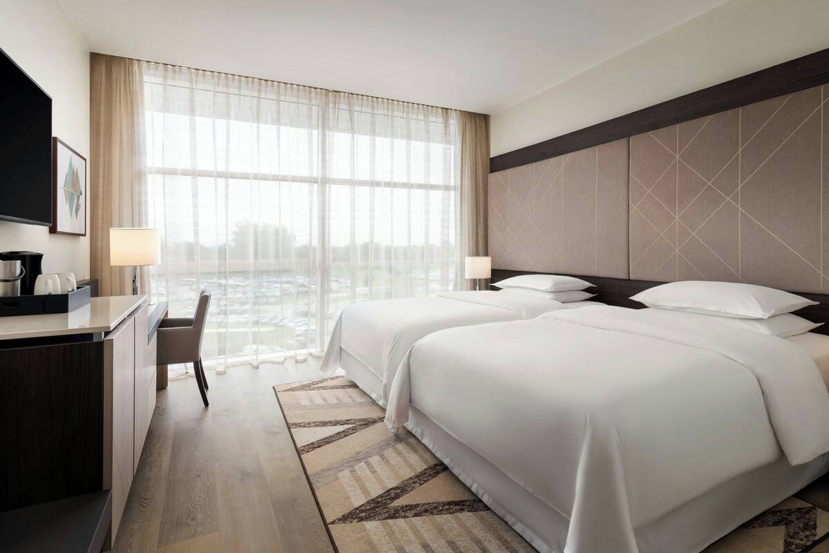 Ein Hotelzimmer mit einem großen Bett, einem Schreibtisch und einem Sessel. Ein Fenster mit leichtem Vorhang bietet einen Ausblick nach draußen.