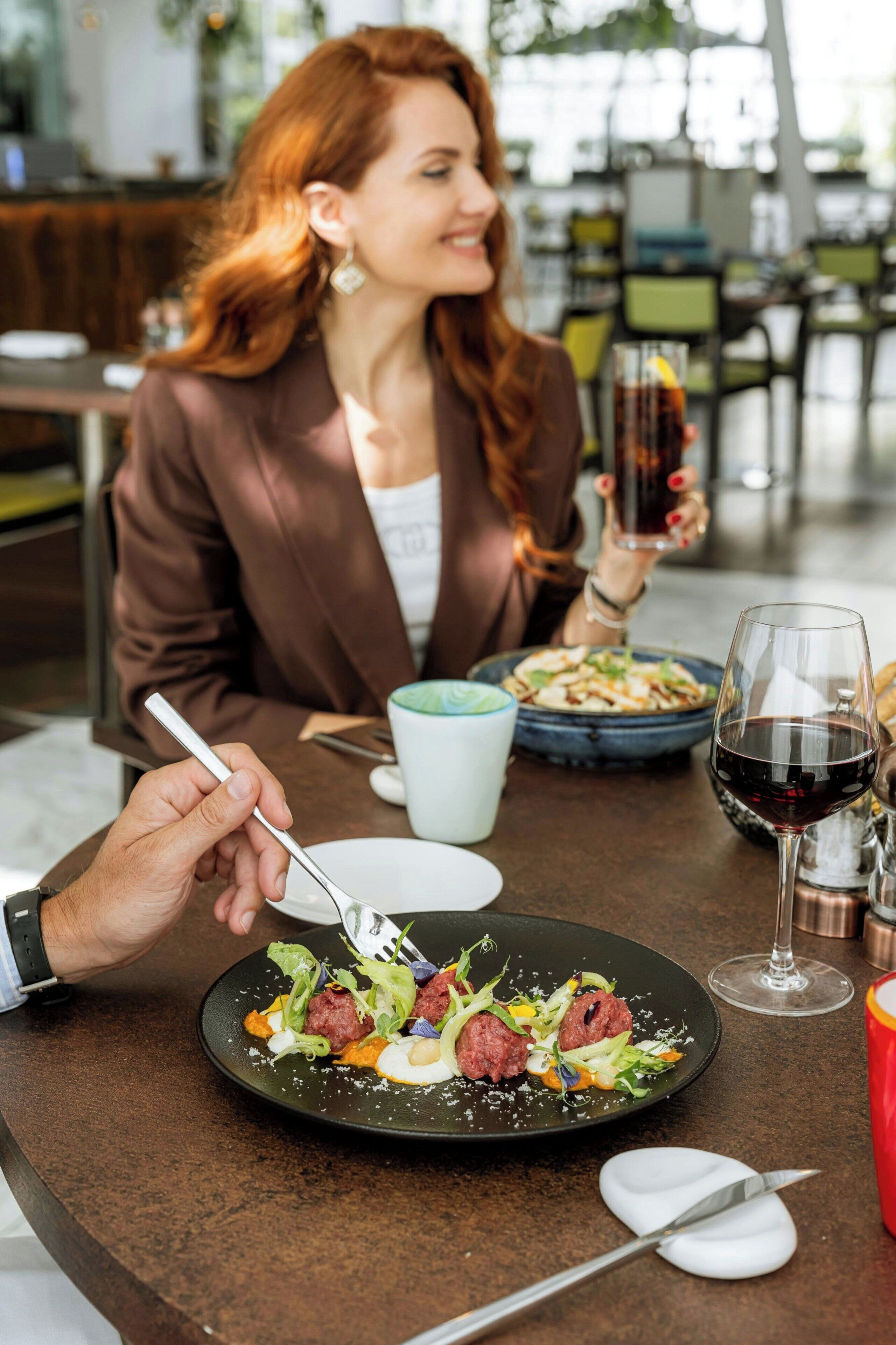 A dining table with a plate of food, a glass of red wine, and a person holding a fork.