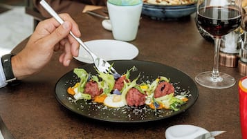 A dining table with a plate of food, a glass of red wine, and a person holding a fork.