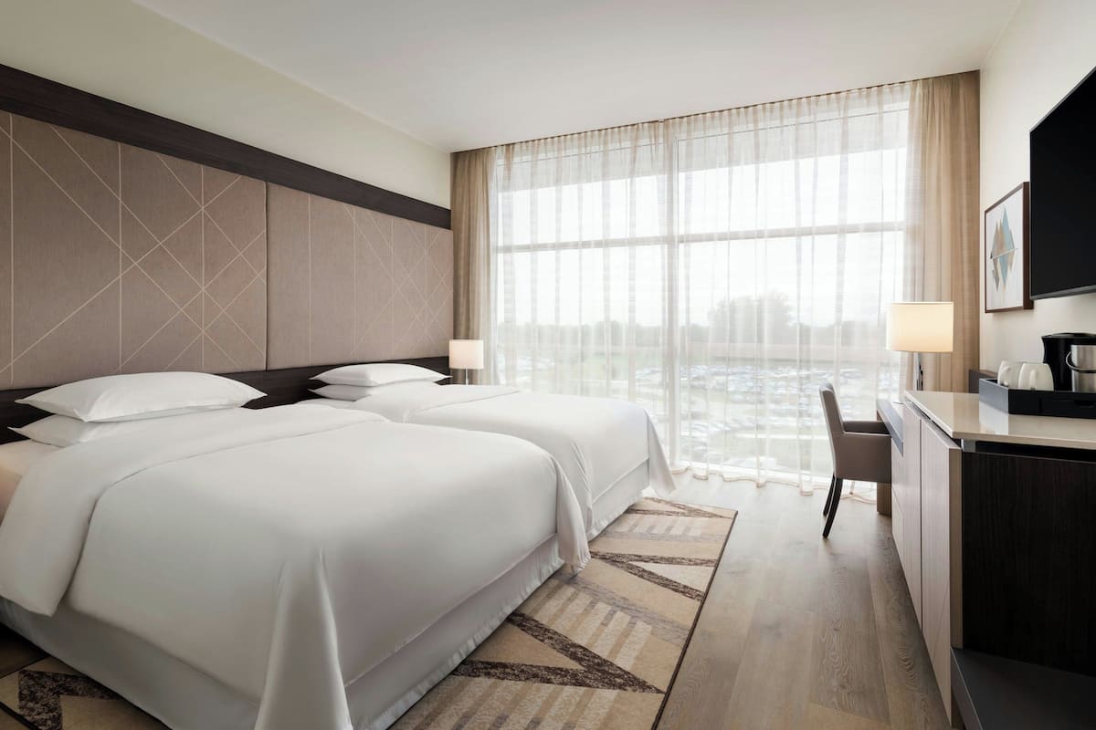 Ein Hotelzimmer mit zwei Betten, einem Schreibtisch, einem Stuhl und einem großen Fenster mit transparenten Vorhängen.