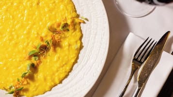 Un piatto di cibo giallo guarnito con erbe aromatiche, accompagnato da forchetta e coltello su un tovagliolo, con un bicchiere di vino rosso sullo sfondo.