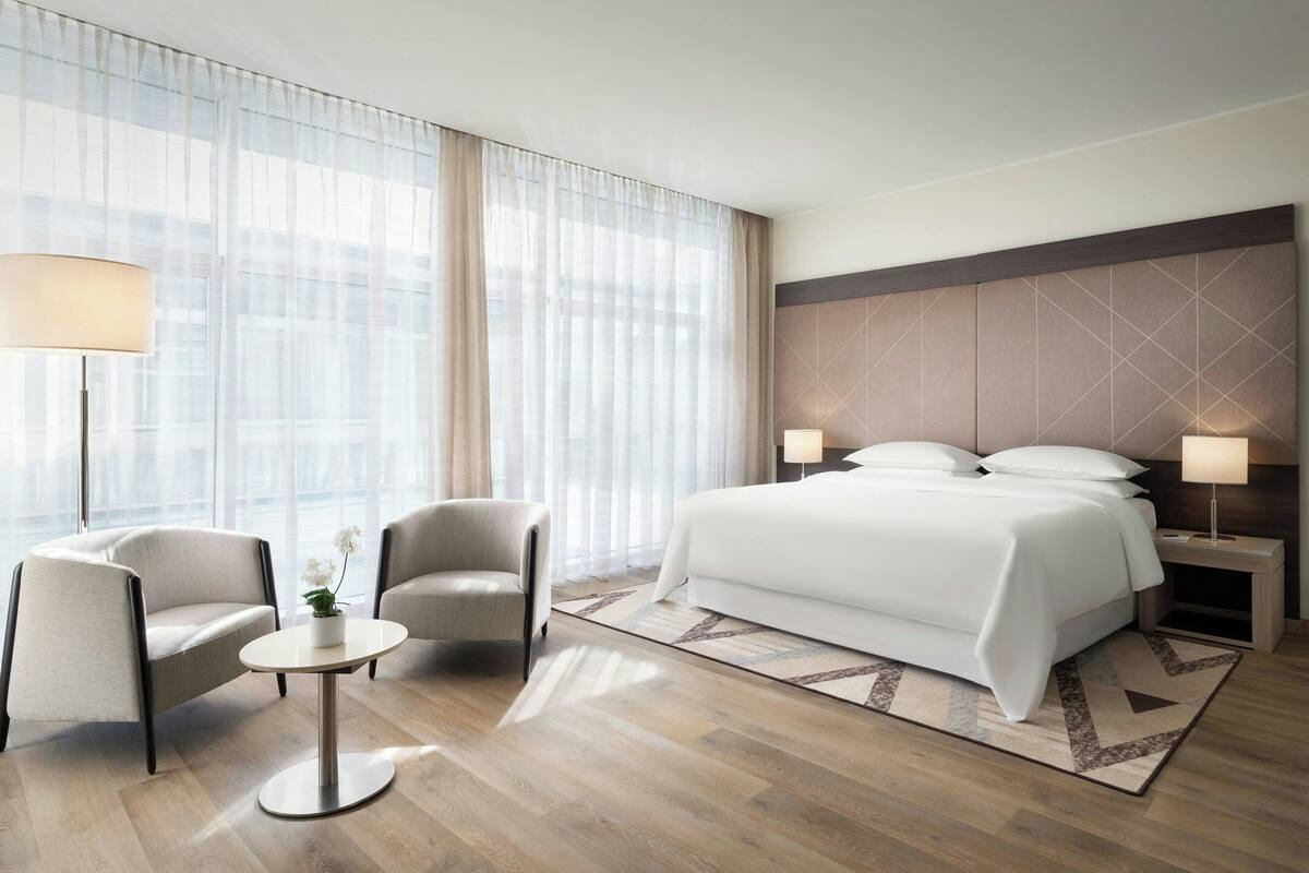 Ein modernes Hotelzimmer mit einem großen Bett, zwei Sesseln, einem kleinen Tisch und einem Nachttisch mit Lampen.