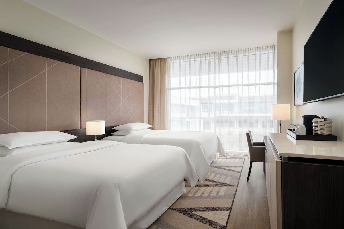 Ein Hotelzimmer mit zwei Betten, einem Schreibtisch, einem Stuhl, einem Fernseher und einem großen Fenster mit Vorhängen.