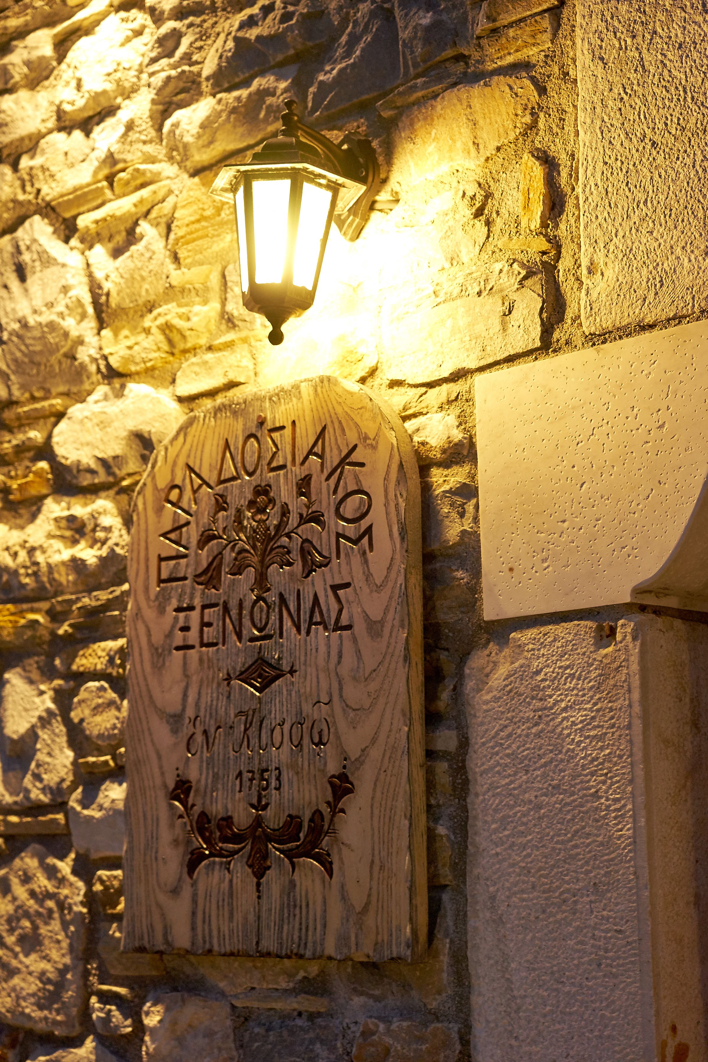 Un muro de piedra con una placa de madera que muestra texto grabado y un emblema decorativo, iluminado por un farol colgante.