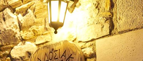 Un muro de piedra con una placa de madera que muestra texto grabado y un emblema decorativo, iluminado por un farol colgante.