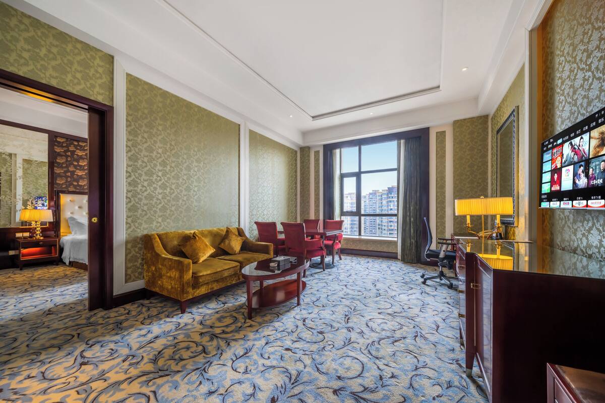 Ruime hotelkamer met een groot raam, een comfortabele gouden bank, een houten salontafel en een aan de muur bevestigde televisie.