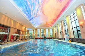 Ein großes Hallenbad mit Deckenprojektion, großen Fenstern und Sitzbereichen mit Sonnenschirmen.