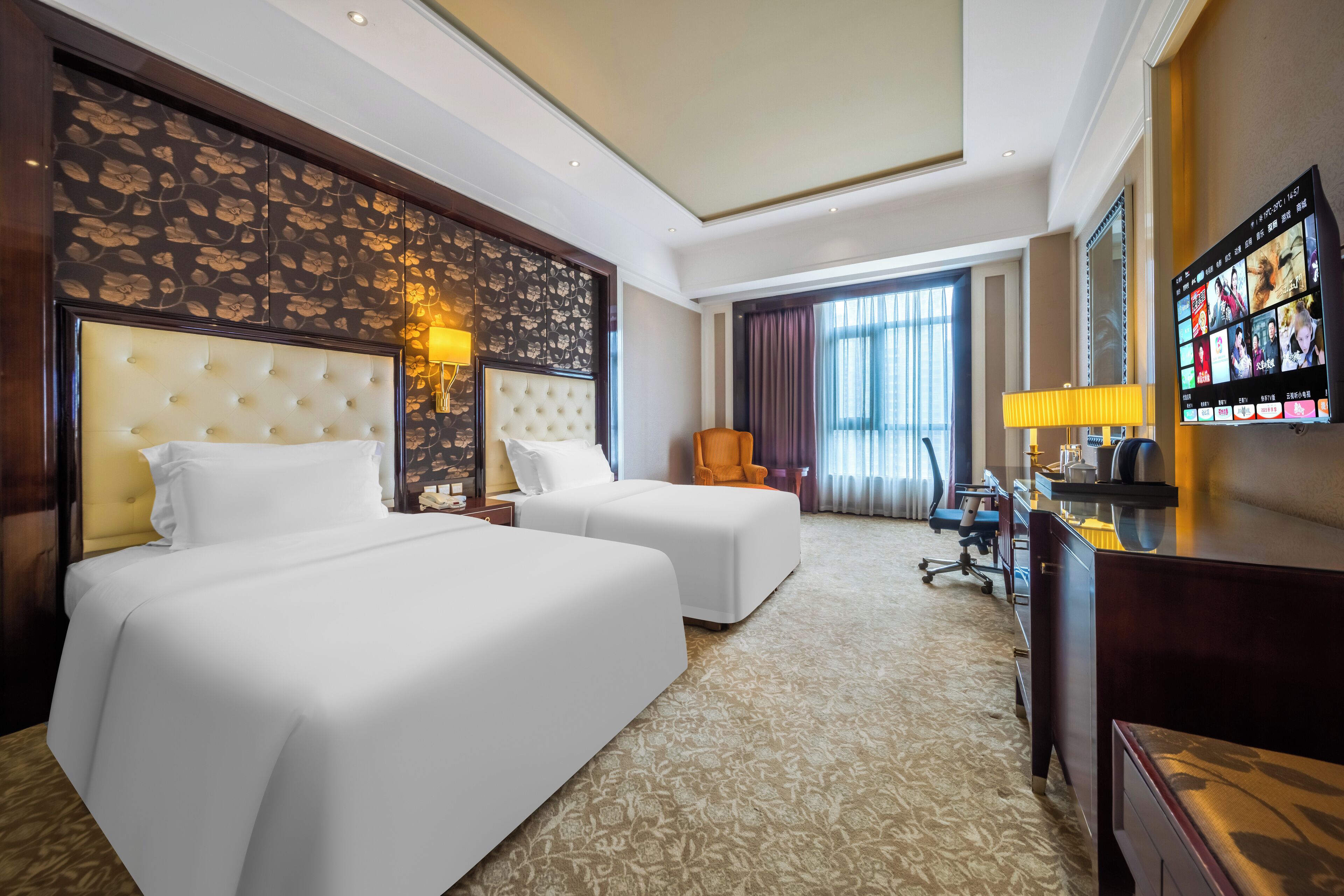 A hotel room with two beds, a desk, a chair, a TV, and a large window with curtains.