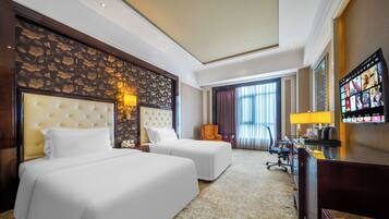 A hotel room with two beds, a desk, a chair, a TV, and a large window with curtains.