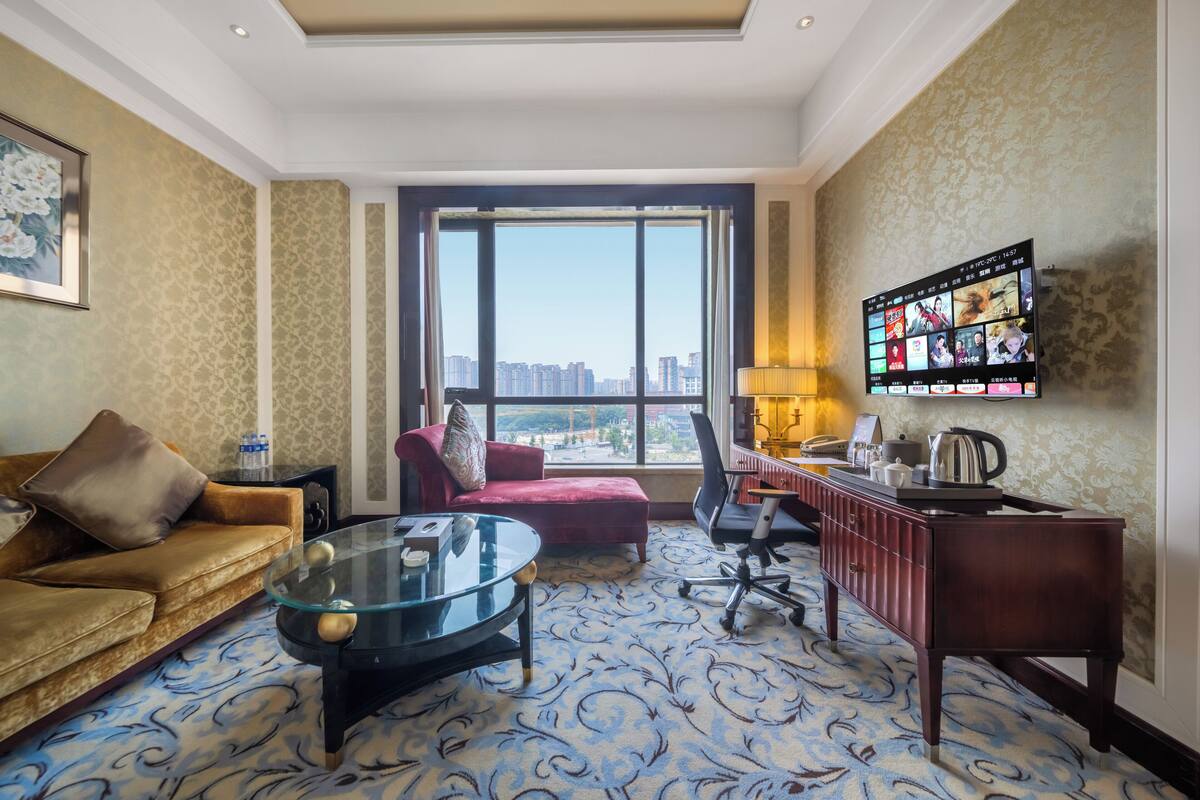 Een hotelkamer met een groot raam, een bank, een bureau en een aan de muur bevestigde televisie.