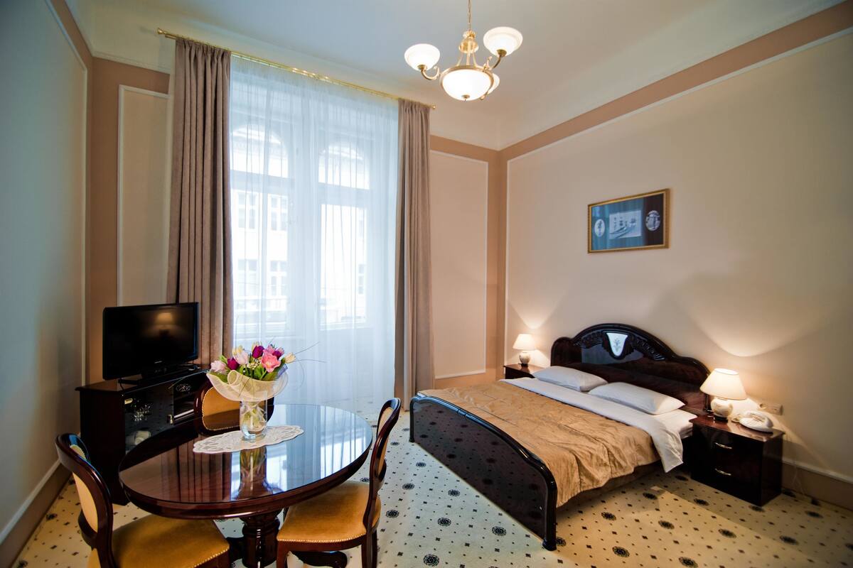 Una habitación de hotel con una cama, mesita de noche con lámparas, televisión, mesa de comedor con sillas y un jarrón de flores.