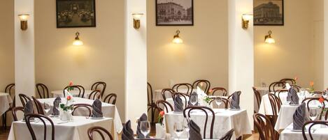 A dining area with tables set for service, framed pictures on the walls, and wall-mounted lighting.
