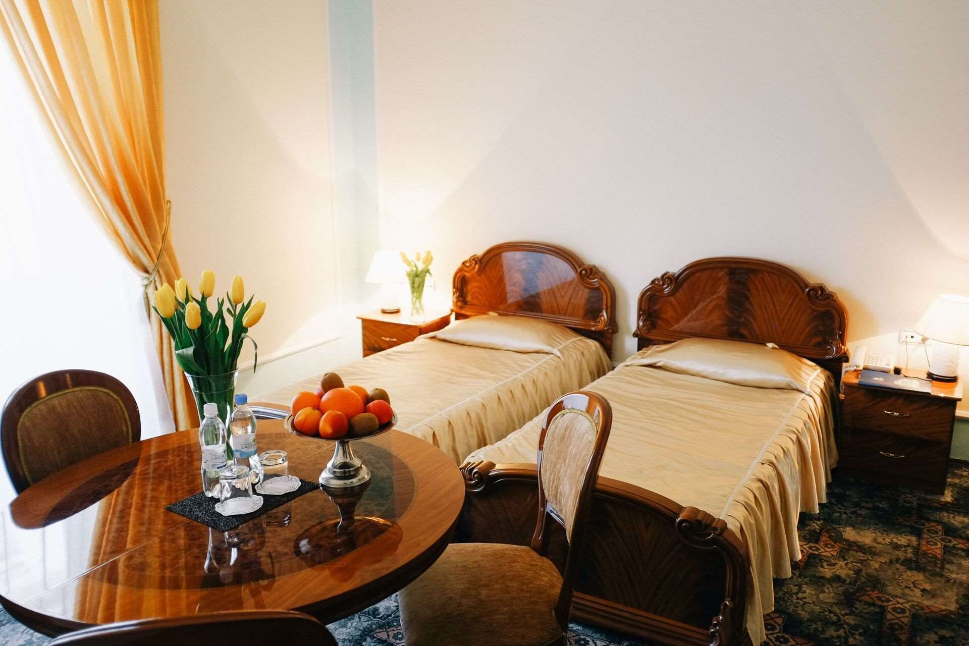 A hotel room with two single beds, a wooden table with a vase of flowers, and a small round table with a fruit bowl.