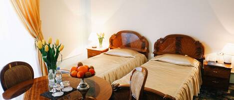 A hotel room with two single beds, a wooden table with a vase of flowers, and a small round table with a fruit bowl.