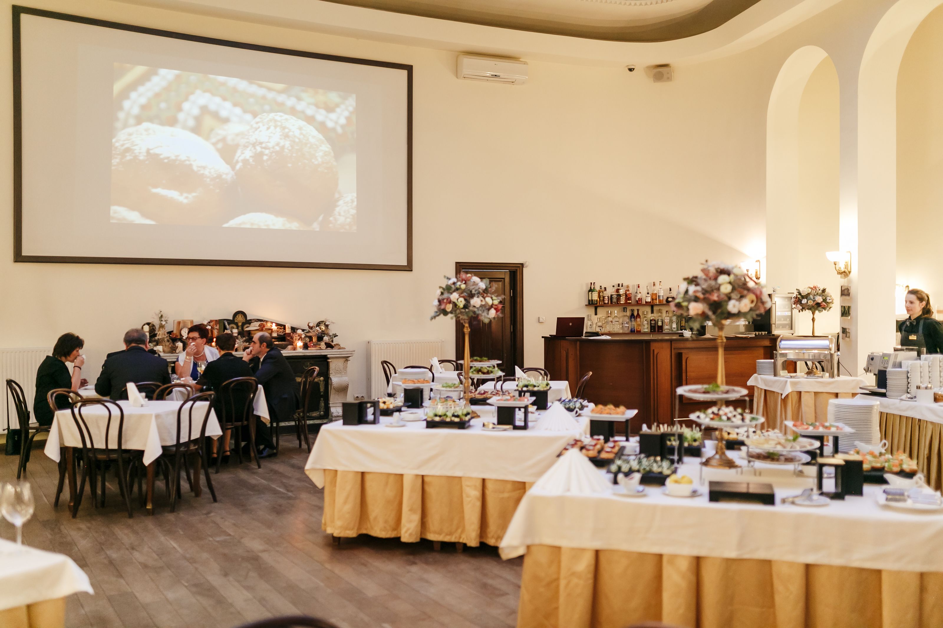 A banquet hall with tables set for a meal, a projection screen, and a bar area with bottles.
