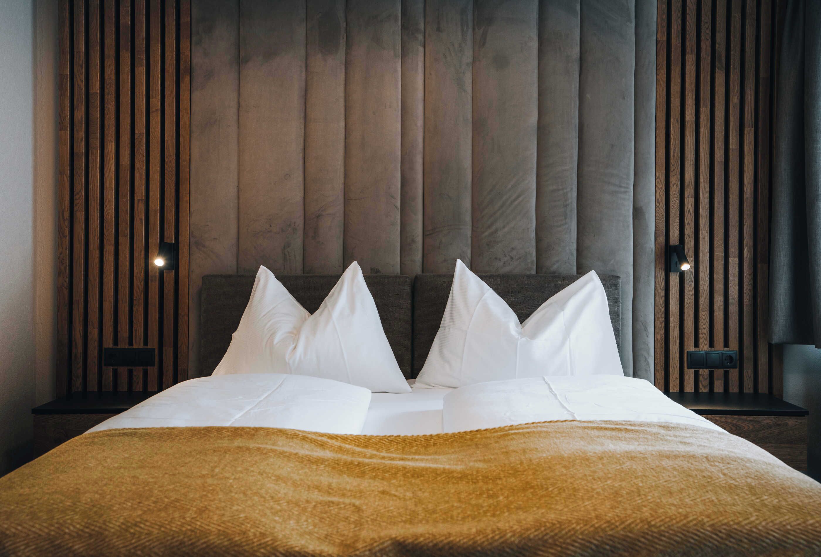 A neatly made bed with white linens and a brown throw blanket, set against a backdrop of vertical wooden slats.