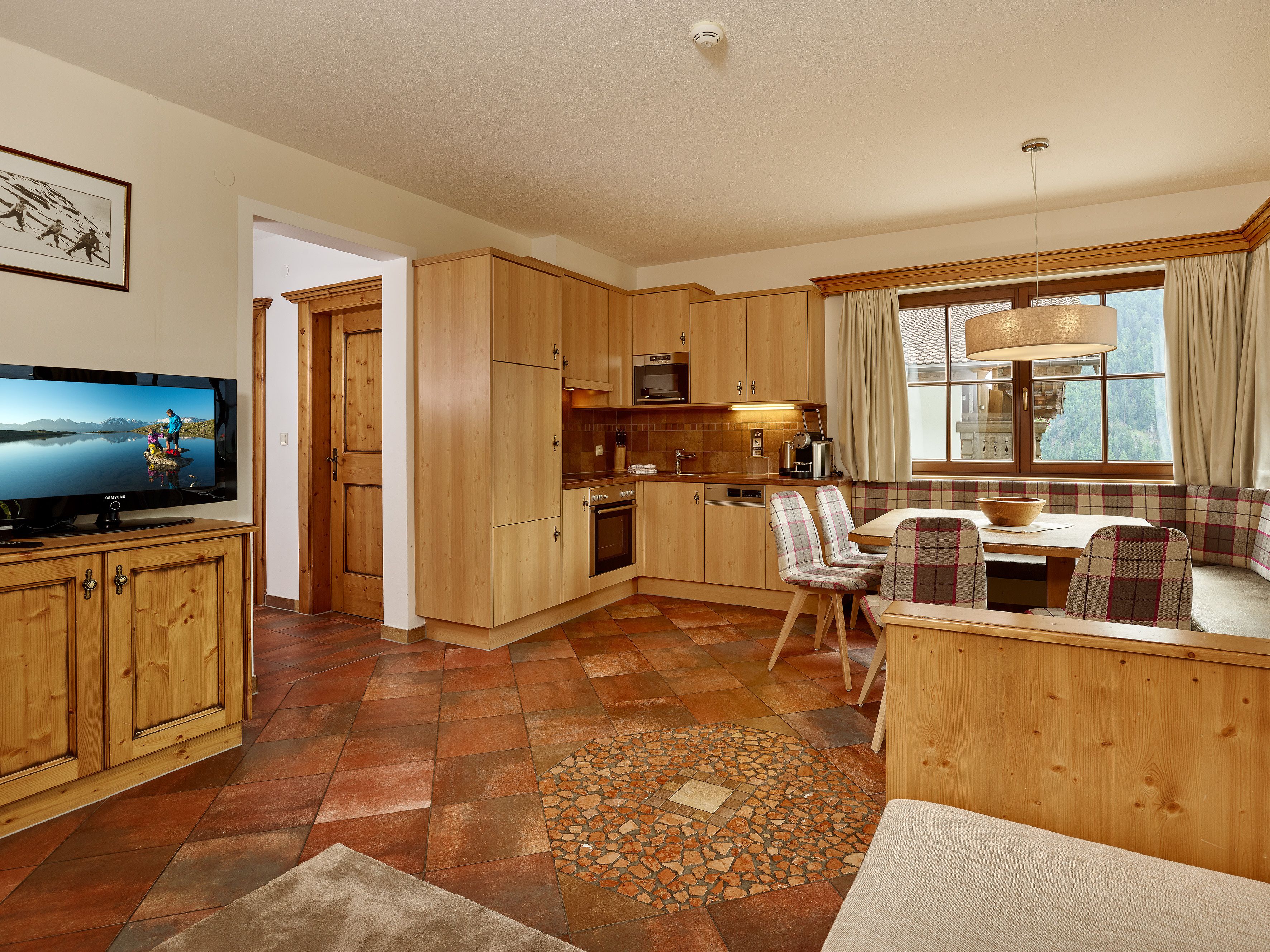 A cozy kitchen with wooden cabinets, a dining area with a bench, and a television displaying a scenic image.