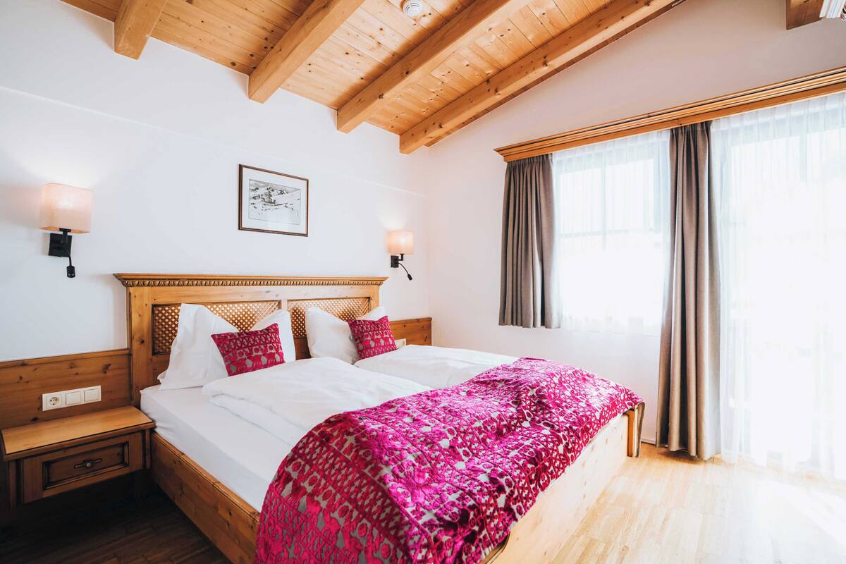 A bedroom with a wooden bed, two bedside tables, a headboard, a window with curtains, and wall-mounted lamps.