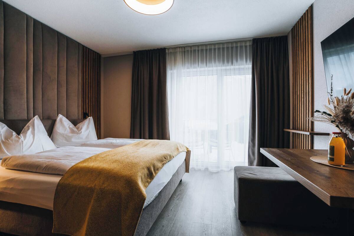 A modern hotel room with a large bed, a wooden desk, and a view of the city through sheer curtains.