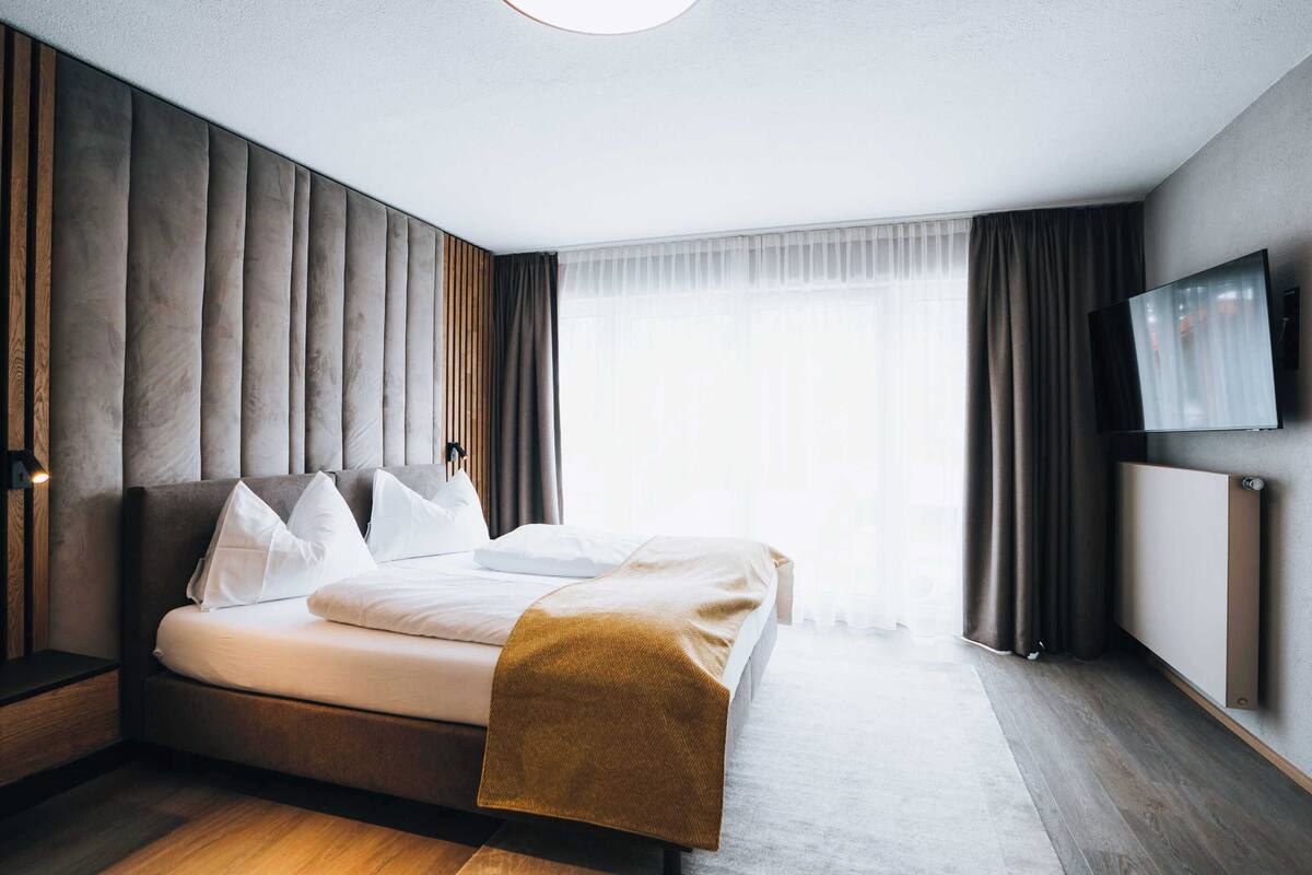 A modern hotel room with a large bed, a flat-screen TV, and a large window with curtains.