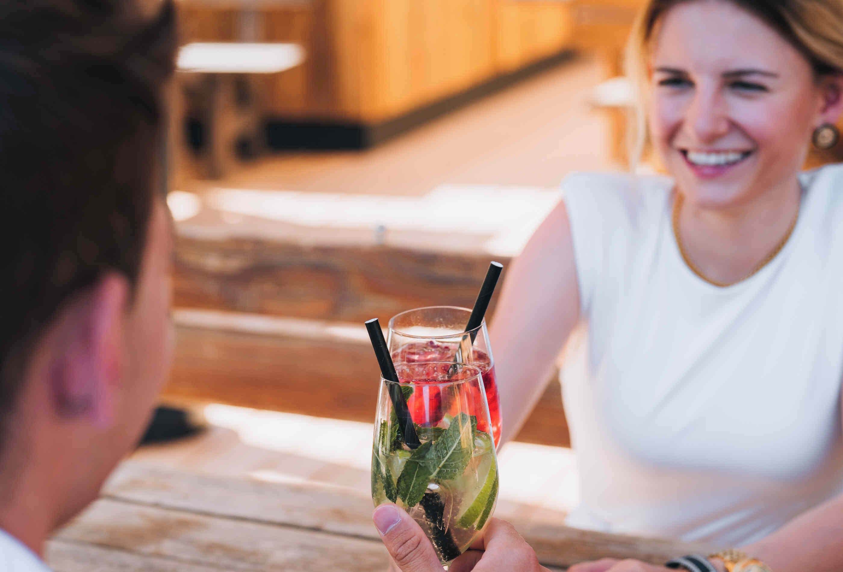 A person holding a drink with a straw and lime slice, sitting at a wooden table in a restaurant.