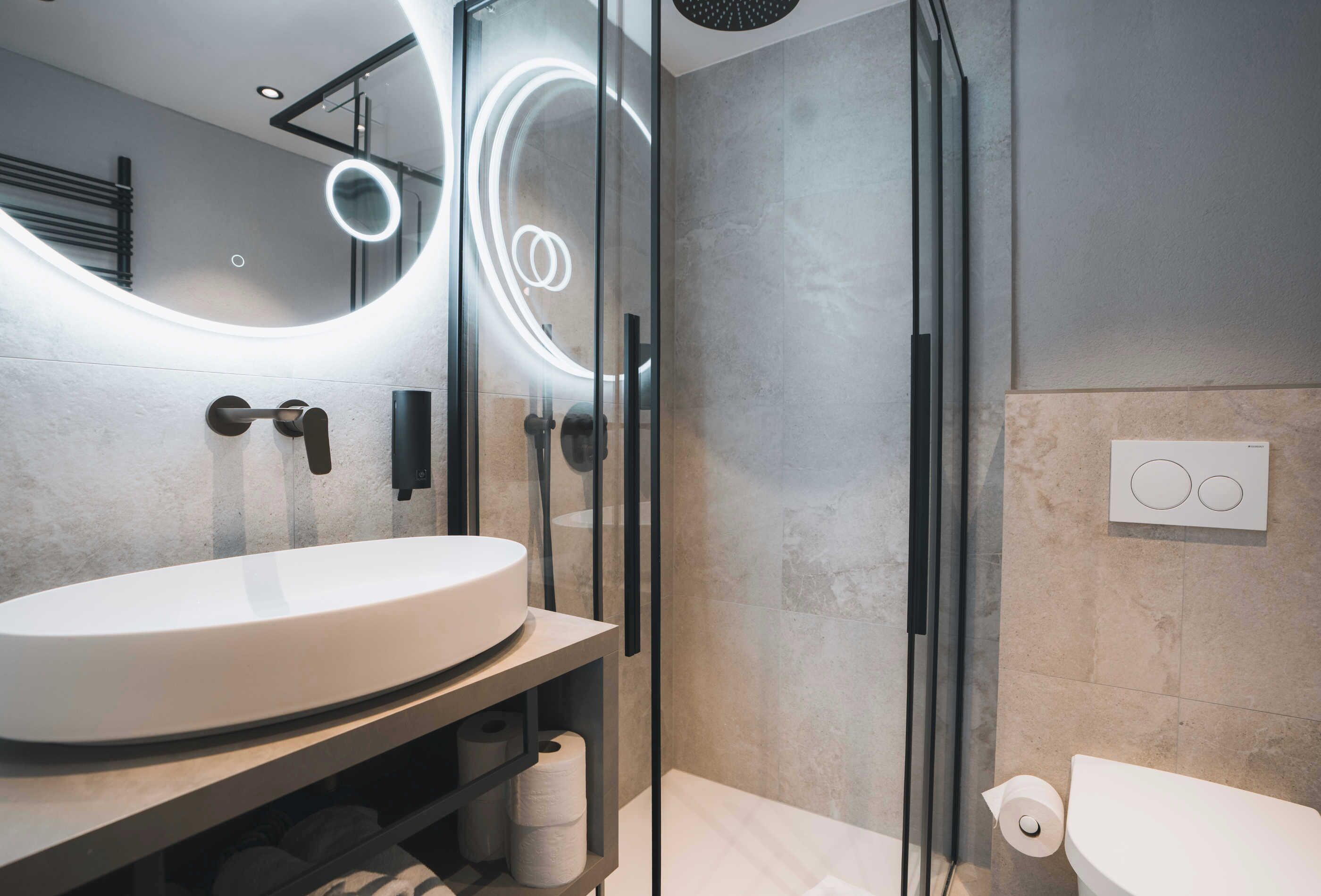 Un bagno moderno con uno specchio rotondo, un lavandino bianco e una cabina doccia in vetro.